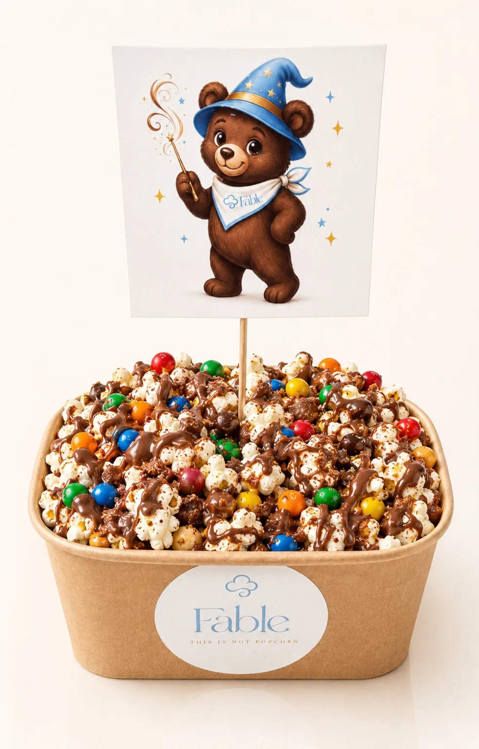 A kraft Fable tub of popcorn with a dark drizzle and colourful candy-coated pieces, topped with an illustration card of a bear in a blue wizard hat.