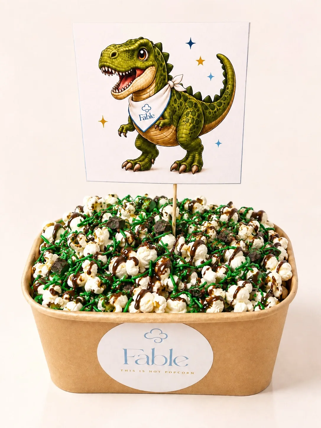 A kraft Fable tub of popcorn with a dark drizzle and bright green sprinkles, topped with a friendly green dinosaur illustration card on a stick.