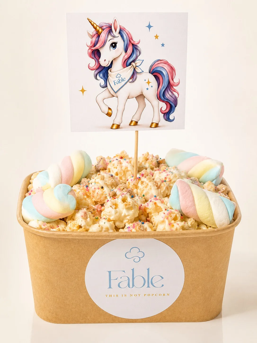 A kraft Fable tub of drizzled popcorn with rainbow sprinkles and pastel twisted marshmallows in the corners, topped with a unicorn illustration card on a stick.